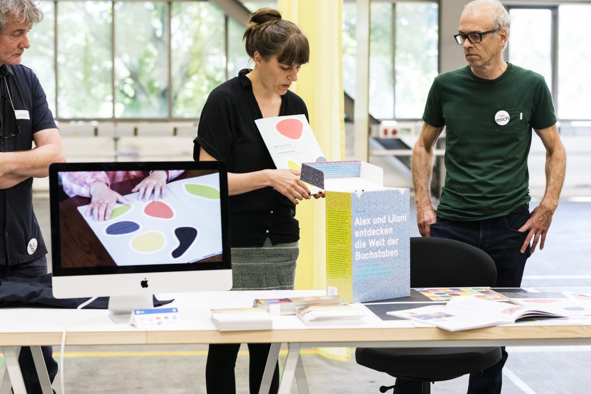 Andréas, Fabienne und Martin präsentieren die Hefte. Vor ihnen steht ein Tisch mit den ausgelegten Heften und ein Screen mit den Heften in Gebrauch von Kinderhänden.